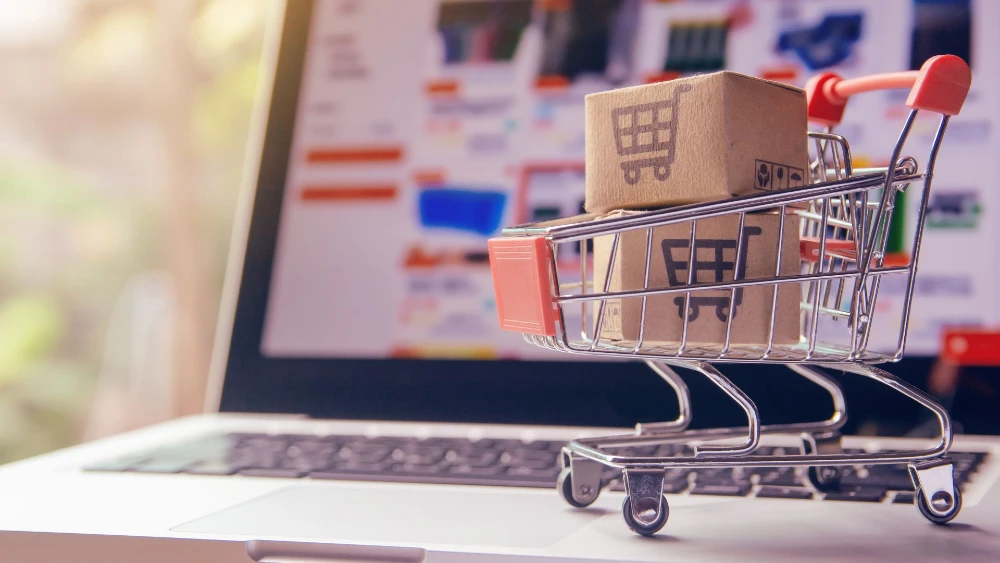 A miniature shopping cart holding two small cardboard boxes sits on a laptop keyboard, with an online store website blurred in the background, symbolizing e-commerce and online shopping.