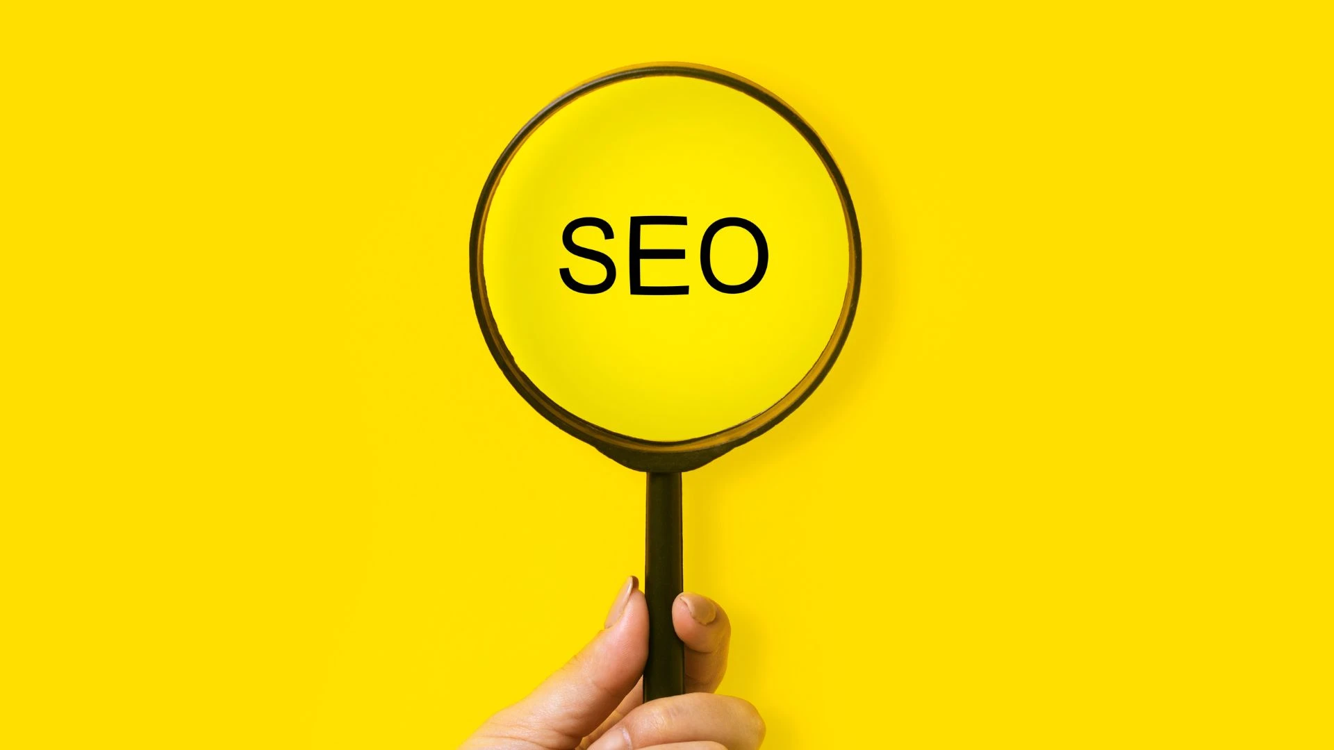 A magnifying glass held by a hand highlighting the word 'SEO' against a bright yellow background, symbolizing the concept of search engine optimization and digital visibility.