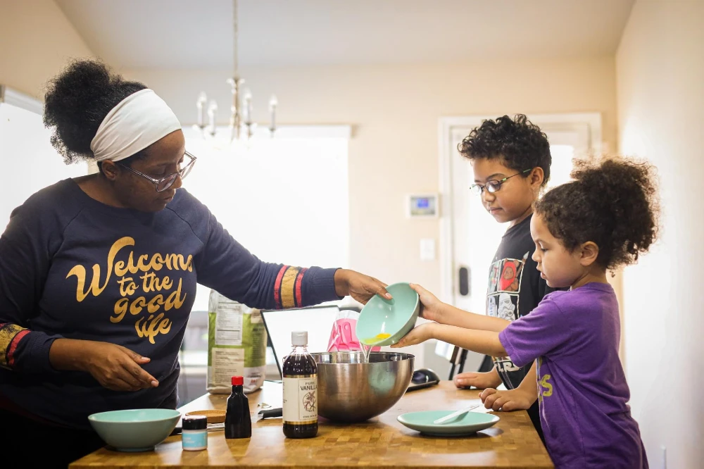 Parent cooking with two children at a kitchen counter, pouring ingredients into a mixing bowl during homeschool activity