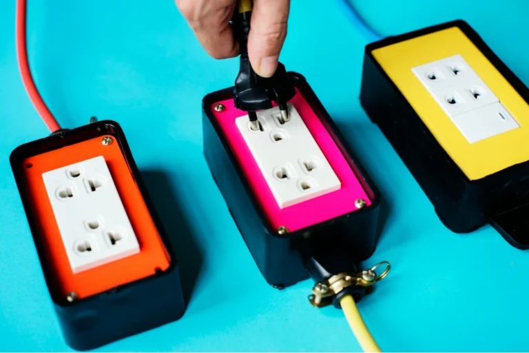 3 brightly colored outlet boxes, one orange, one hot pink, and one highlighter yellow - all in a row with a sea blue background. a hand is seen plugging a cord into the middle box.