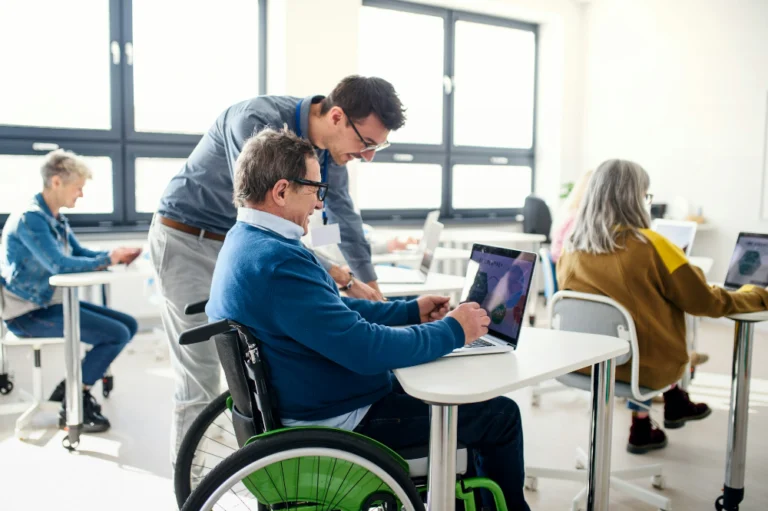This photo depicts several people sitting at desks in an all white room, working at laptops. The man in the foreground is sitting at his desk in a wheelchair and another man is leaning in looking at his laptop.