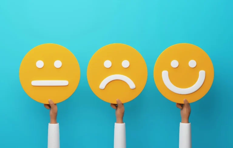 There are 3 different arms accending from the bottom of screen in a white long sleeved T-shirt. Each hand is holding a vibrant yellow "smiley" face against a sea blue background. The first hand's smiley face has a straight line for a mouth, showing a neutral expression. The middle hand's smiley is actually a frown, and the right hand's smiley is a true smiling face.